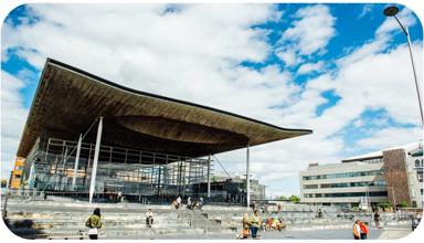 A photograph of the front of the Senedd building in Cardiff Bay. The building is comprised of grey bricks, and some white beams are visible. There is a sign that reads ‘Welsh Parliament’ in the back right hand side. In the foreground, there are pedestrians passing by. Some people are sitting on the steps outside the Senedd building.