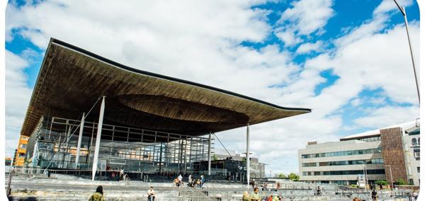 A photograph of the front of the Senedd building in Cardiff Bay. The building is comprised of grey bricks, and some white beams are visible. There is a sign that reads ‘Welsh Parliament’ in the back right hand side. In the foreground, there are pedestrians passing by. Some people are sitting on the steps outside the Senedd building.