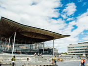 A photograph of the front of the Senedd building in Cardiff Bay. The building is comprised of grey bricks, and some white beams are visible. There is a sign that reads ‘Welsh Parliament’ in the back right hand side. In the foreground, there are pedestrians passing by. Some people are sitting on the steps outside the Senedd building.